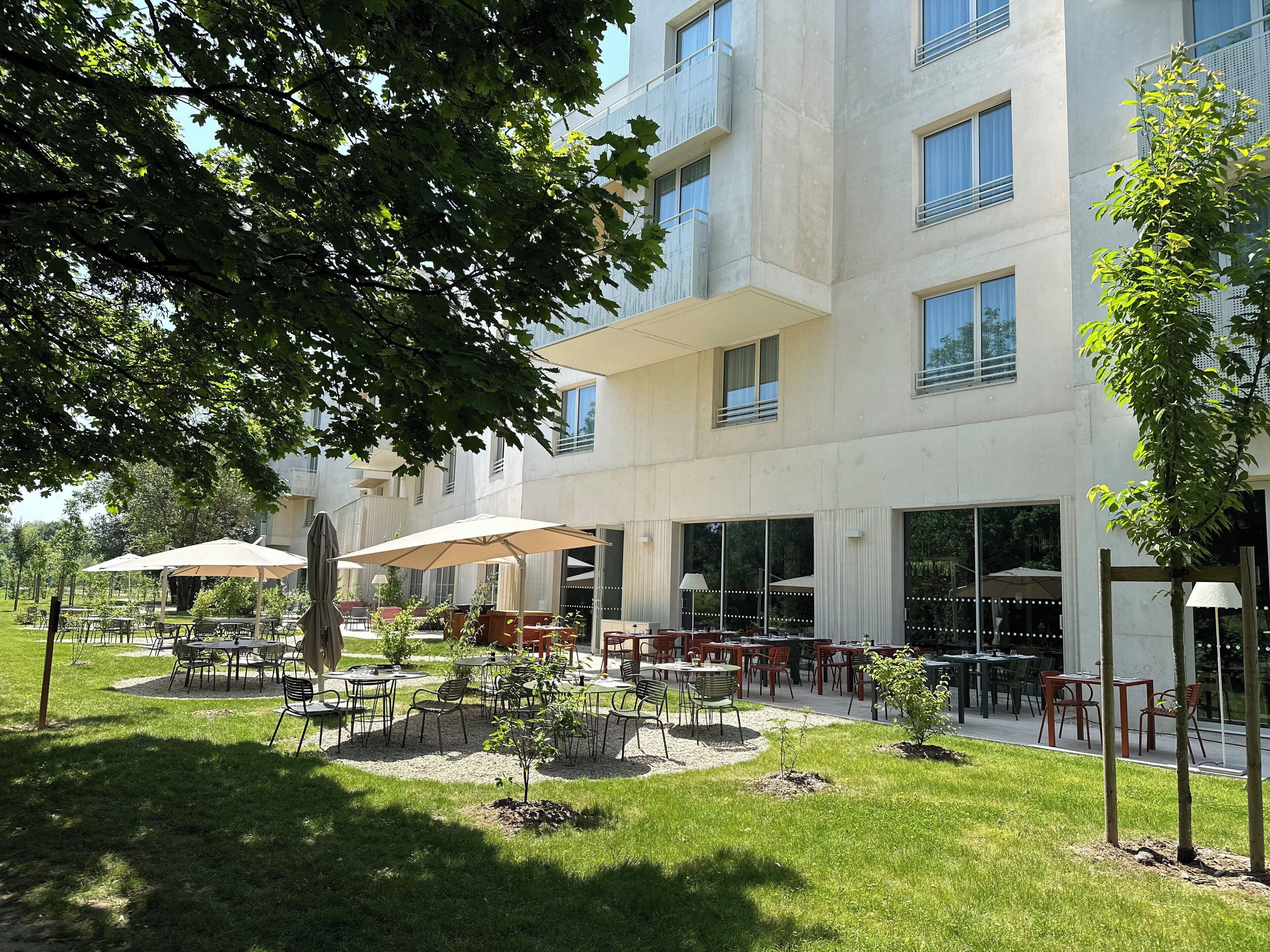 An outdoor patio area with tables and chairs, surrounded by greenery and a modern building with large windows.