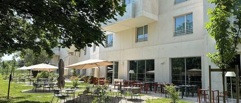 An outdoor patio area with tables and chairs, surrounded by greenery and a modern building with large windows.