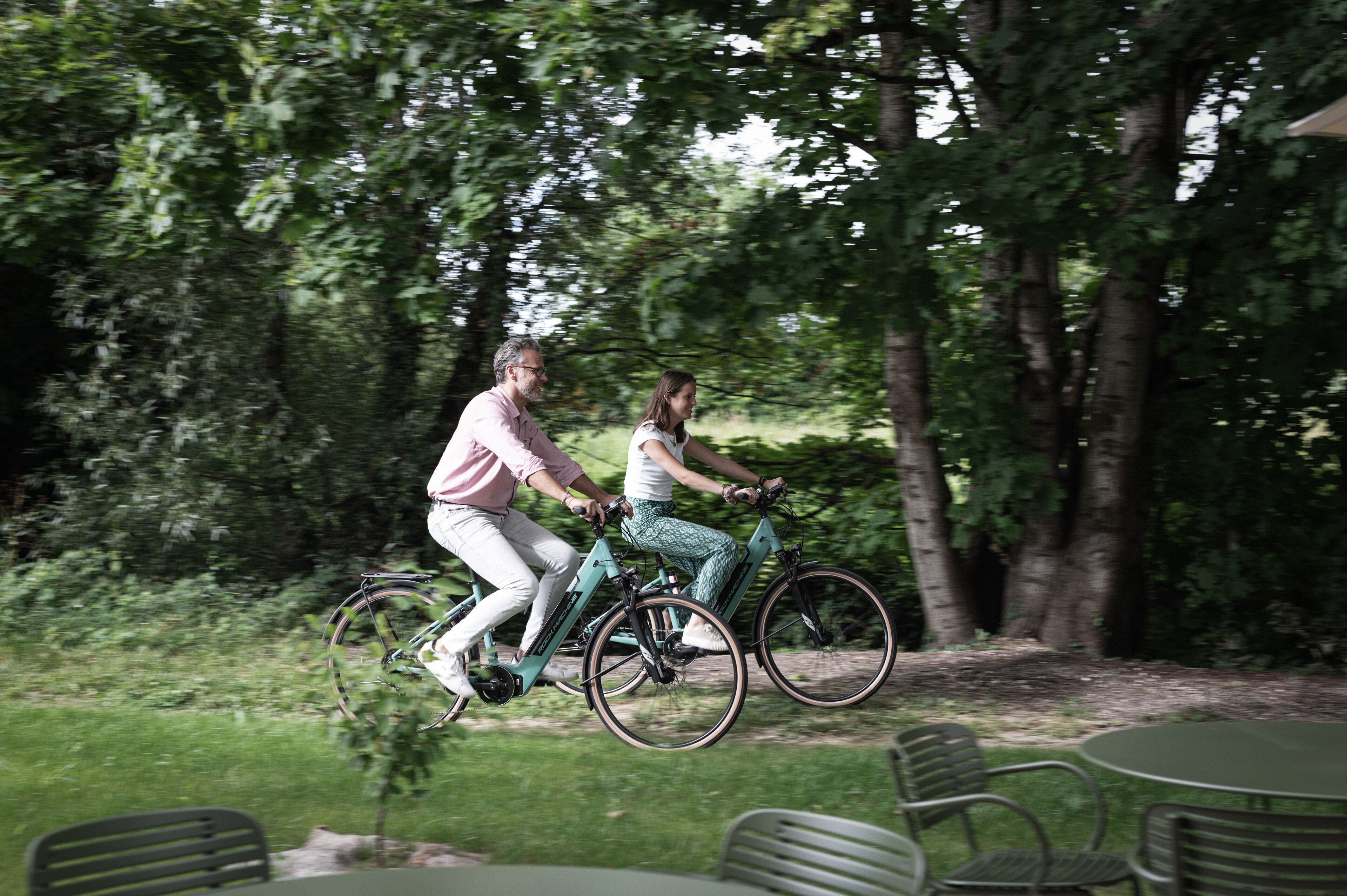 Deux personnes font du vélo d’appartement dans un parc, près d’arbres et de tables et chaises installées à l’extérieur.