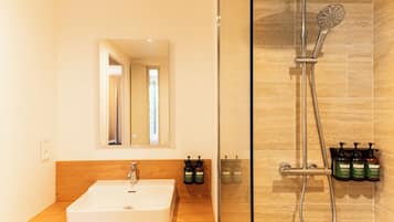 Un baño con lavamanos blanco, espejo y ducha con cabezal de ducha manual y botellas de champú.