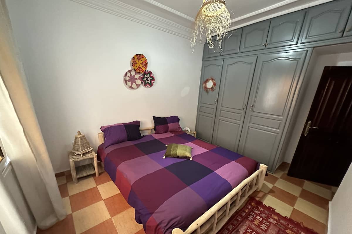 Un dormitorio con suelo a cuadros, una cama con colcha morada y azul, cabecero de madera, mesita de noche, cesta tejida y un reloj colgado en la pared.