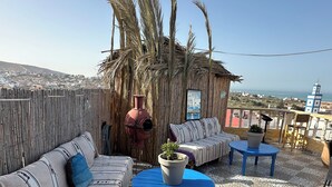 A terrace with a thatched-roof hut, a patterned sofa, a blue table, and a potted plant. - DAR SANAE SURF (Aourir)