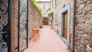 A narrow alleyway with a brick floor, stone walls, and a wooden door. There is a small table with chairs outside a building. - Le Dimore di Borgo Tessile - Feronia (Orvieto)