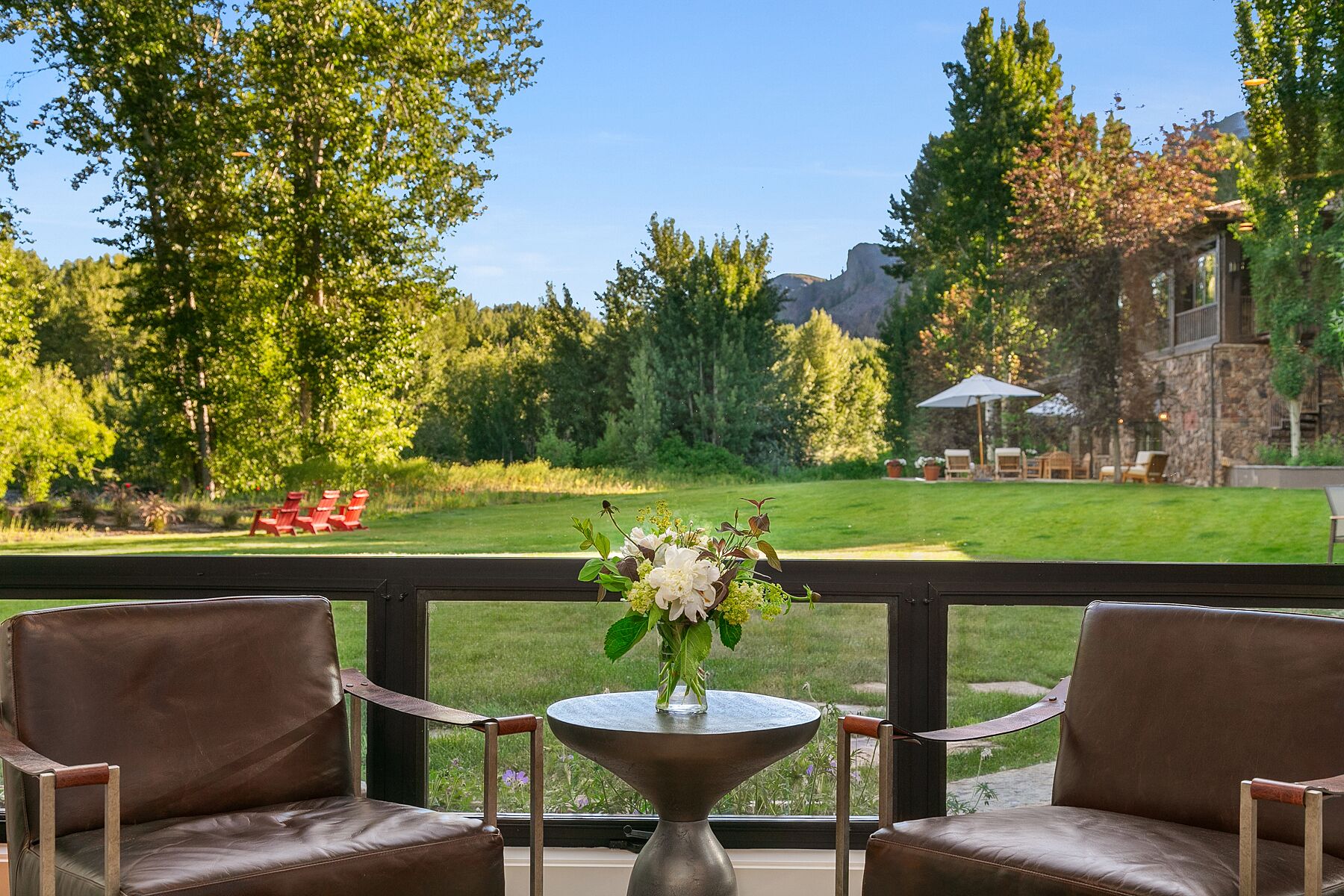 A balcony with two leather chairs, a small round table with a vase of flowers, and a view of a well-maintained lawn and trees.