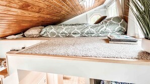 Camera soppalco con soffitto spiovente, un letto con biancheria a motivi, un tavolino e una pianta in vaso.