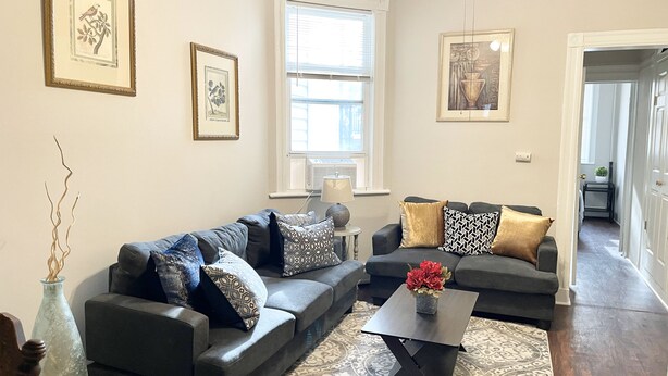 A living room with two sofas, a coffee table, and decorative pillows. There are framed pictures on the walls and a window with blinds.