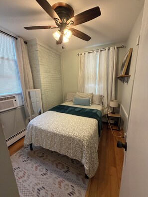Ein Schlafzimmer mit einem Bett, Deckenventilator, einem fenster mit Vorhängen, einem Heizkörper und einem an der Wand befestigten Regal.