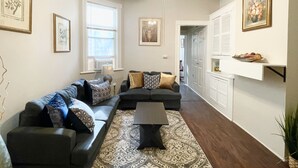 A living room with a sectional sofa, a coffee table, and a ceiling fan. There are framed pictures on the walls and a window with blinds.