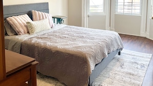 A bedroom with a large bed, a headboard, a wall-mounted artwork, and a window with blinds.