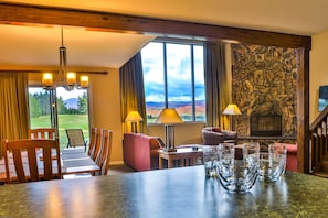 A room with a stone fireplace, a dining table with chairs, a sofa, and a view of a golf course. - Marcy Lodge u4 - Magnificent Mountain Views (Lake Placid)