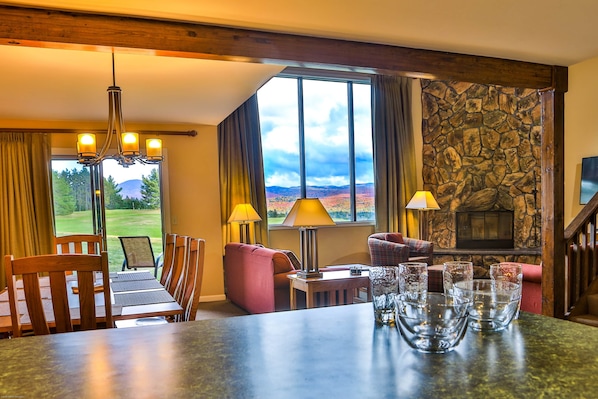 A room with a stone fireplace, a dining table with chairs, a sofa, and a view of a golf course.