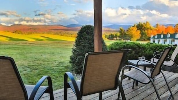 A covered outdoor seating area with chairs and tables, overlooking a scenic landscape with fields and distant hills. - Marcy Lodge u1 - Magnificent Mountain Views! (Lake Placid)