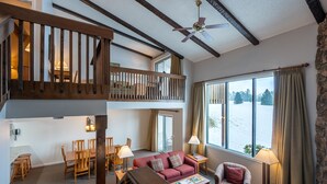 A spacious living area with a high ceiling, wooden beams, a balcony, a dining area, and a large window with a view of trees.
