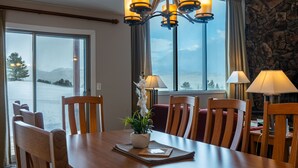 A dining area with a wooden table, chairs, and a chandelier. There is a view of a body of water through large windows.