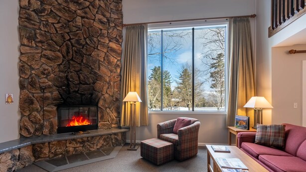 A cozy living room with a stone fireplace, a large window with curtains, a plaid armchair, a red sofa, and a wooden coffee table.