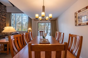 Un espace repas comprenant une longue table en bois, des chaises, un lustre et une vue dégagée grâce à de grandes fenêtres.