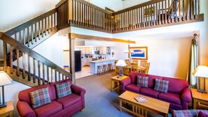 A spacious living area with a wooden staircase, a kitchen in the background, and a red sofa with plaid cushions.