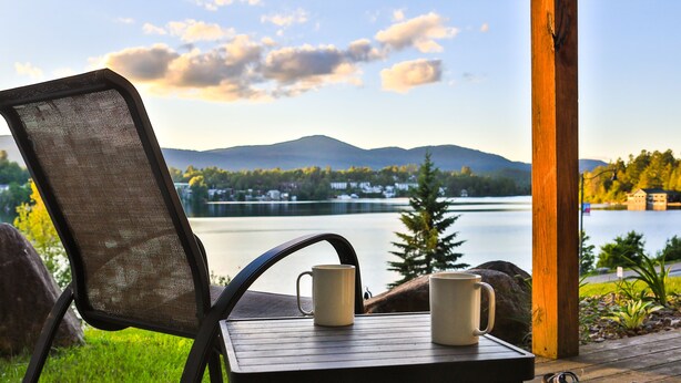 A wooden deck with a table and a chair overlooking a lake, with mountains in the background.