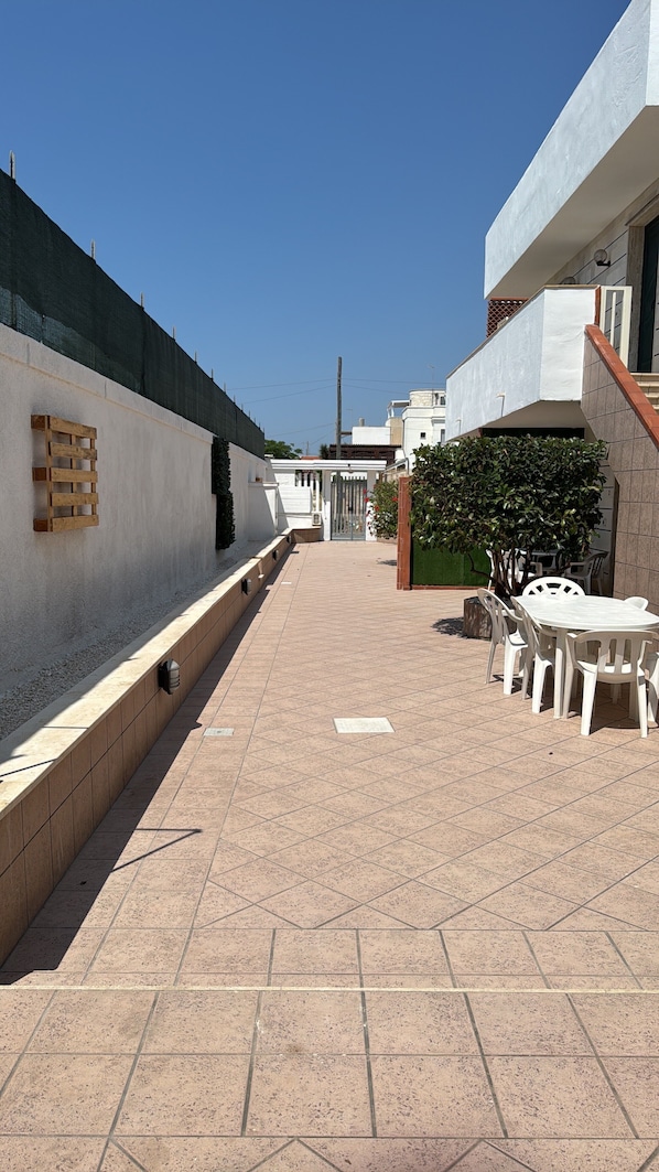 A courtyard with a tiled floor, a wooden bench, a white table with chairs, and a green hedge.