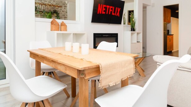 A modern living room with a wooden dining table, white chairs, a TV displaying 'NETFLIX', and a fireplace. - Steps from Grandvalira  Cozy Ski Haven+Parking

 (Soldeu)