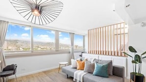 A modern living room with a grey sofa, a small round table, and a large window offering a city view. - NEWLY Remodeled | Upscale Design | Central Waikiki (Honolulu)