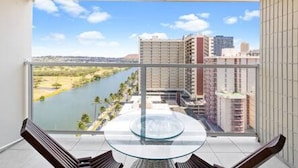 A balcony with a glass table and chairs, overlooking a river and buildings. - NEWLY Remodeled | Upscale Design | Central Waikiki (Honolulu)