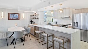 A modern kitchen with a central island, bar stools, a dining table with chairs, stainless steel appliances, and pendant lighting.