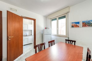 A dining area with a wooden table and chairs, a kitchen with white cabinets, a refrigerator, and a window with blinds.