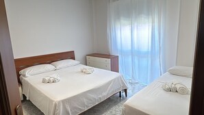 Ein Hotelzimmer mit zwei Betten, einem Holzkopfteil, einem Nachttisch, einer weißen Kommode und einem Fenster mit durchsichtigen Vorhängen.