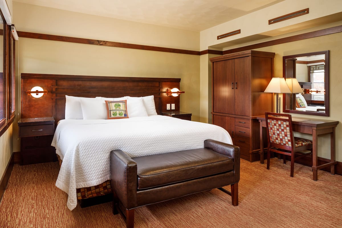 A hotel room with a large bed, a wooden desk, a chair, a bench, and a wardrobe.