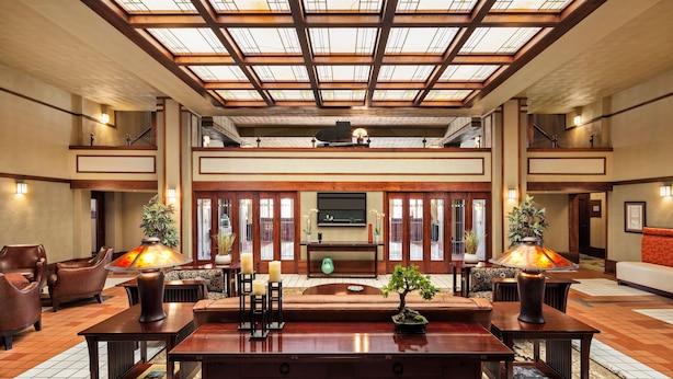 A spacious lobby with a high ceiling, wooden beams, and a central seating area with a sofa and coffee table. There are two lamps on either side of the seating, a small table with a vase, and a potted plant. - The Historic Park Inn Hotel (Mason City)