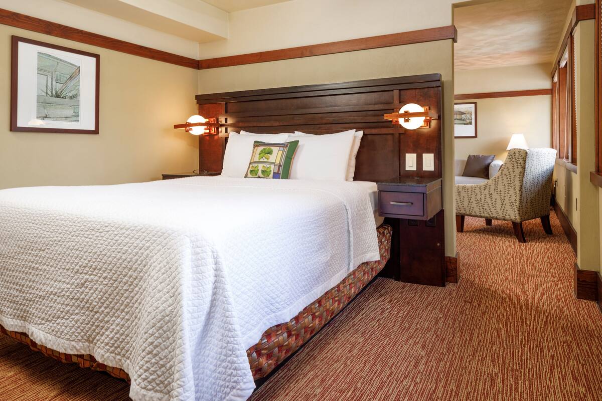 A hotel room with a large bed, a wooden headboard, two bedside lamps, a chair, and a carpeted floor.