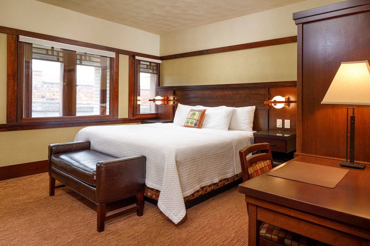 A hotel room with a large bed, a wooden headboard, a desk, and a chair. There are two bedside lamps and a window with blinds.