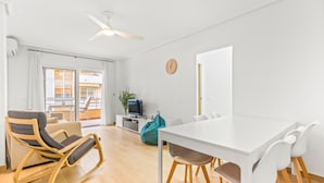A modern living room with a white dining table, a rocking chair, a TV, and a balcony with curtains. - Apartment 'Moriones Playa' with Balcony, Wi-Fi and Air Conditioning (Torrevieja)