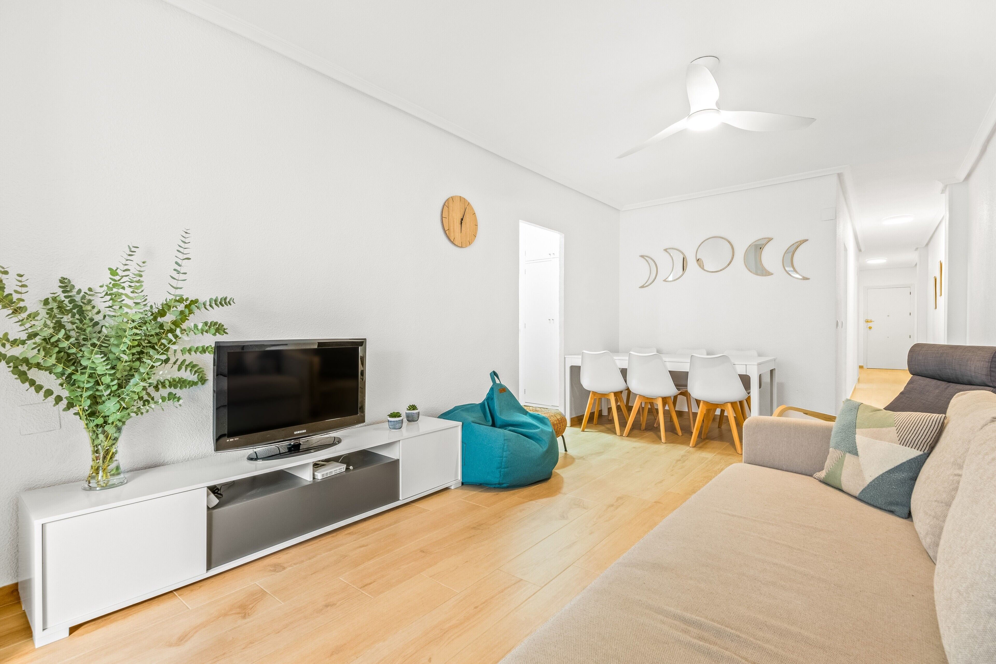Een moderne woonkamer met een flatscreen-tv, een witte bank, een blauwgroene relaxfauteuil, een eethoek met een witte tafel en stoelen, en een plafondventilator.
