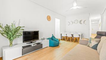Een moderne woonkamer met een flatscreen-tv, een witte bank, een blauwgroene relaxfauteuil, een eethoek met een witte tafel en stoelen, en een plafondventilator.