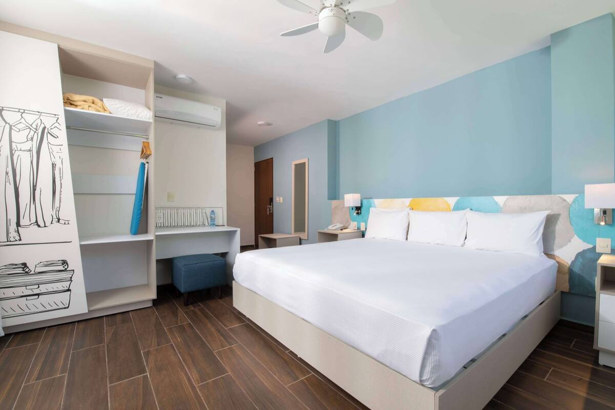 Een moderne hotelkamer met een groot bed, een garderobe met daaraan kleding, een bureau en een plafondventilator.