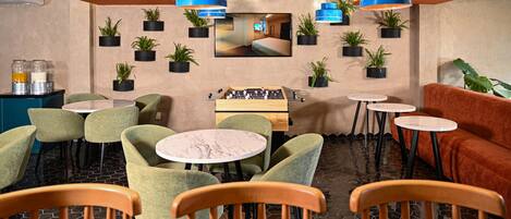 A cozy indoor seating area with round tables and chairs, a foosball table, and potted plants on the walls.