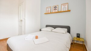 Una cama bien hecha con sábanas y almohadas blancas, una mesita de noche con una lámpara y un estante en la pared con tres obras de arte enmarcadas.