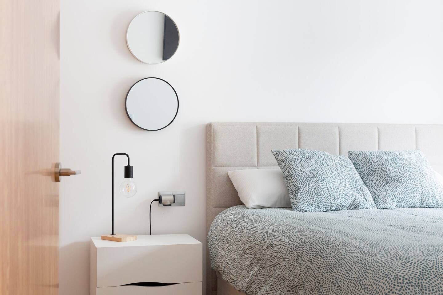 Un dormitorio con cama, dos almohadas, mesita de noche con lámpara y espejo en la pared.