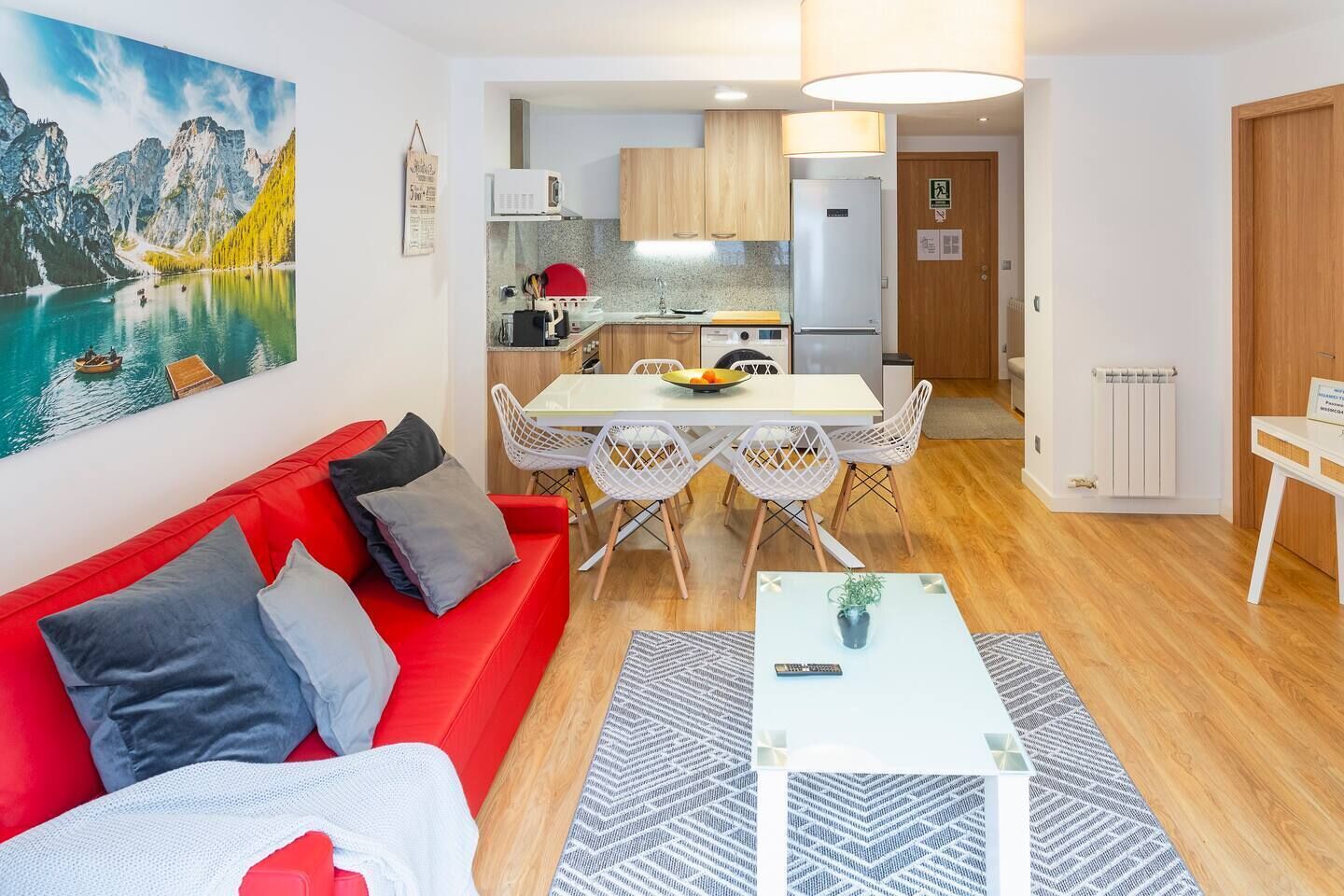 Una sala de estar moderna con un sofá rojo, una mesa de comedor con sillas blancas y una cocina con armarios de madera y un refrigerador.