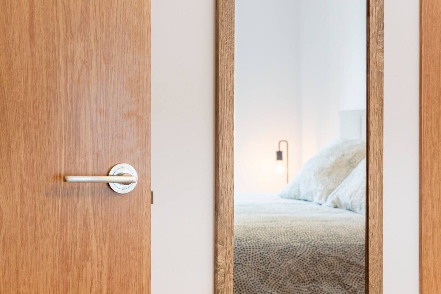 A wooden door with a modern handle and a mirror reflecting a bedroom with a bed and a wall-mounted light.
