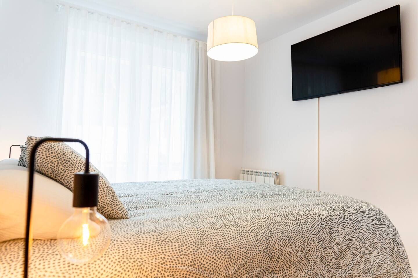 Un dormitorio con cama, una lámpara, televisor en la pared y una ventana con cortinas.