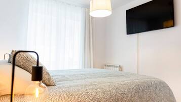 Ein Schlafzimmer mit einem Bett, einer Lampe, einem an der Wand befestigten Fernseher und einem Fenster mit Vorhängen.