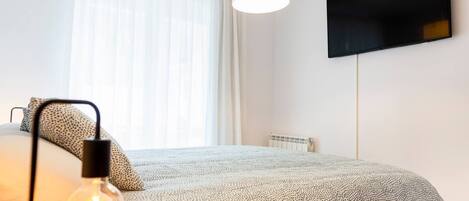 Un dormitorio con cama, una lámpara, televisor en la pared y una ventana con cortinas.