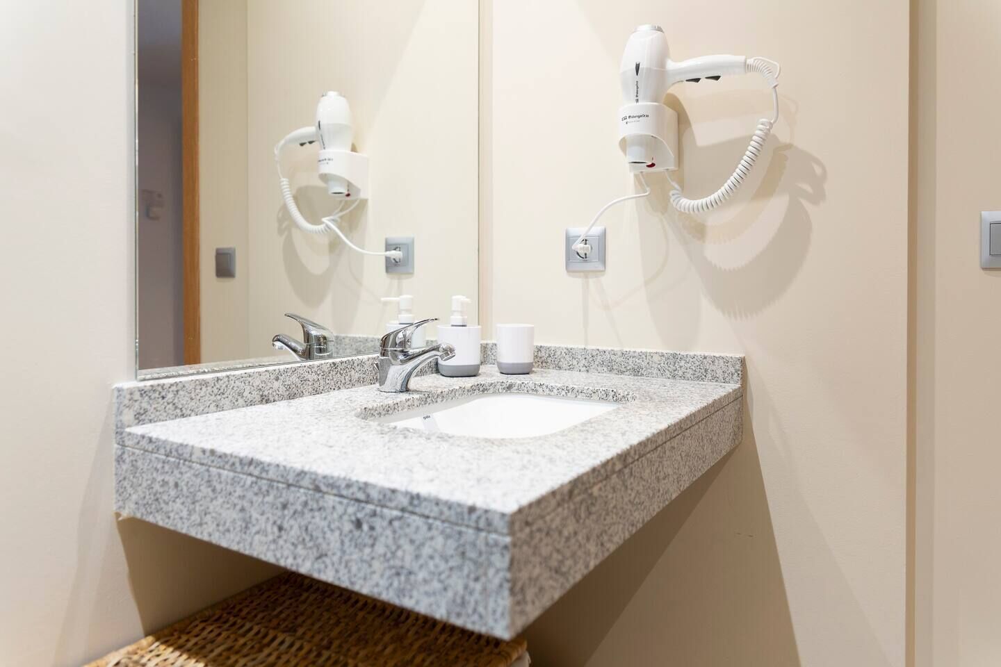 A bathroom with a granite sink, two hair dryers mounted on the wall, and a mirror above the sink.