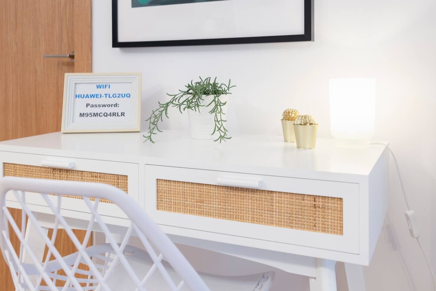 Un escritorio blanco con un cartel del código de acceso WiFi enmarcado, una planta pequeña y dos jarrones decorativos.