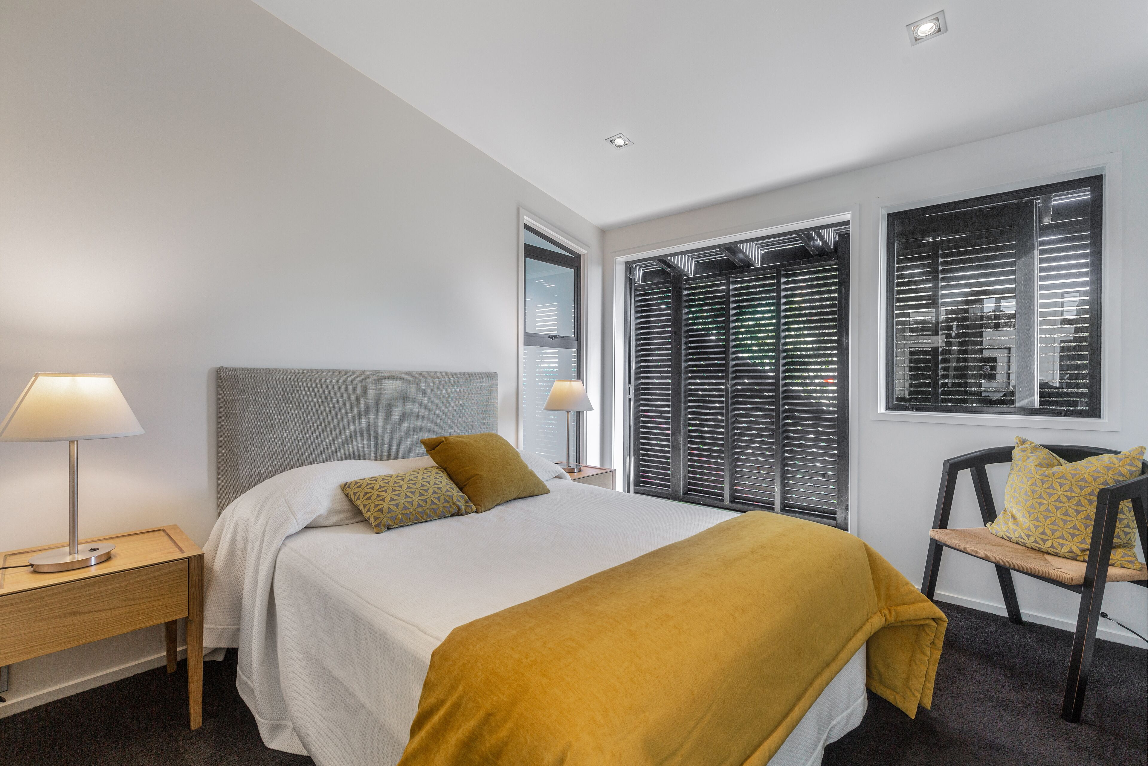Une chambre d’hôtel moderne avec un grand lit, une table de chevet avec une lampe, une chaise et une fenêtre avec des stores.