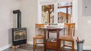 Un comedor con una mesa redonda y cuatro sillas, un hogar de leña y una ventana con vista a una construcción de madera en el exterior.