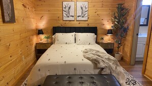 Un dormitorio acogedor con una pared de madera, una cama con colcha blanca decorada con motivos de árboles y huellas de patas, dos mesitas de noche con lámparas, un banco negro y una alfombra estampada.